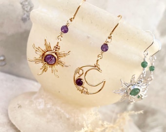 May include: Gold-toned and silver-toned celestial earrings. One pair features a sun design with a purple gemstone center. Another pair has a crescent moon with purple gemstones. The third pair has a sun design with a green gemstone center.