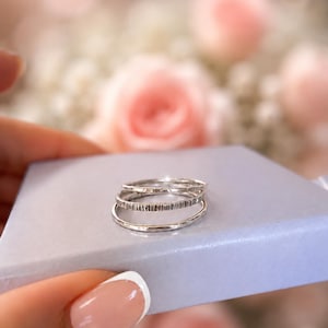 Può includere: Un set di tre anelli in argento impilati su una scatola azzurra. Gli anelli presentano texture distinte, tra cui una fascia liscia, una fascia martellata e una fascia con linee verticali incise, realizzate in metallo lucido.