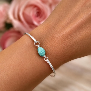 Peut inclure: Un bracelet jonc en argent avec une texture martelée et une pierre turquoise centrale en forme de goutte. La pierre est fixée par un fil d'argent enroulé et reliée aux extrémités du jonc par de petits anneaux en argent, créant un aspect délicat.