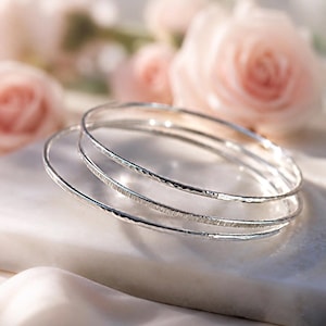 Puede incluir: Un conjunto de tres brazaletes circulares y finos de plata. Cada brazalete presenta una textura de superficie distinta, incluyendo uno con acabado martillado, otro con apariencia cepillada o rayada y uno con superficie metálica lisa y brillante.