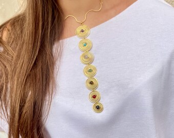May include: A gold-colored necklace featuring a series of spiral-shaped pendants, each adorned with a different colored gemstone. The necklace is worn against a white top, showcasing the jewelry's design and color variations. The gemstones include red, green, blue, and brown.