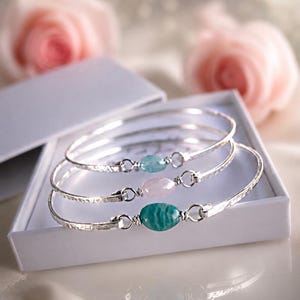 Peut inclure: Trois bracelets rigides en métal argenté martelé, chacun orné d'une pierre précieuse ovale centrée sur le devant. Les pierres sont bleu clair, rose pâle et vert sarcelle. Chaque pierre est fixée au bracelet par un fil métallique argenté.