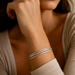Op de afbeelding: Een set van drie dunne, zilverkleurige metalen armbanden gedragen om de pols. Elke armband heeft een glad, afgerond profiel met een subtiele, getextureerde afwerking die het licht opvangt, wat zorgt voor een gestapelde uitstraling op de arm.