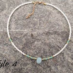Pu&ograve; includere: Una delicata collana di perline bianche con un ciondolo in pietra blu chiaro e dettagli dorati. La collana &egrave; su una superficie grigia testurizzata. Stile 4.