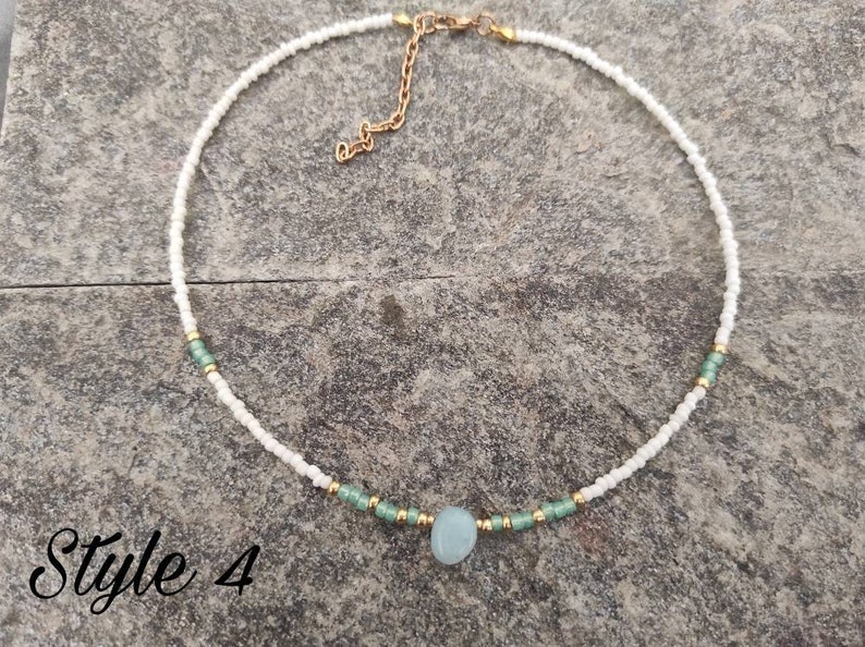 Pu&ograve; includere: Una delicata collana di perline bianche con un ciondolo in pietra blu chiaro e dettagli dorati. La collana &egrave; su una superficie grigia testurizzata. Stile 4.