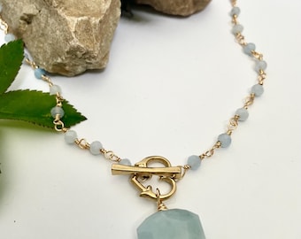 May include: A gold-colored necklace with a toggle clasp and a light blue, faceted gemstone pendant. The necklace features small, light blue beads strung along a gold chain. The pendant is a hexagonal shape. The necklace is displayed on a white surface.