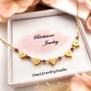 May include: A gold-colored necklace with heart-shaped pendants spelling out "JULIA". The necklace features small, round, red gemstones between the heart pendants. The necklace is presented in a white box with the text "Birthstone Jewelry" and "Duet Jewelry Studio".