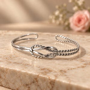 Peut inclure: Un bracelet manchette en argent avec un motif de nœud central. Le bracelet est composé de deux bandes distinctes : l'une avec une finition martelée et texturée, et l'autre avec un aspect torsadé en forme de corde, se rejoignant au nœud central.
