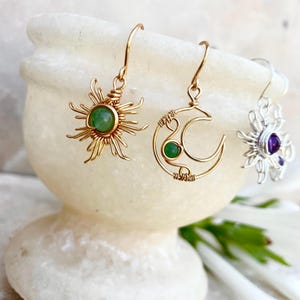 Può includere: Tre paia di orecchini a tema celeste. Un paio presenta un design a forma di sole dorato con un centro in pietra verde. Un altro paio ha una luna crescente dorata con una pietra verde. Il terzo paio è argentato con una pietra viola.