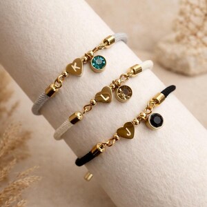 Op de afbeelding: Drie gepersonaliseerde armbanden met goudkleurige hartbedels, initialen en edelsteenaccenten. De armbanden hebben grijze, witte en zwarte koorden. De bedels en accenten zijn goudkleurig.