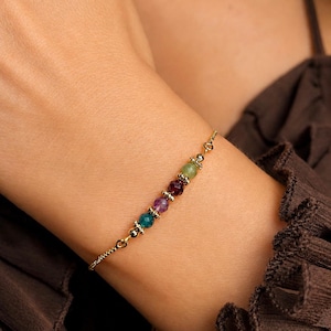 Può includere: Un braccialetto dorato con una serie di perline di pietre preziose colorate. Le perline, nei toni del verde, viola, rosso e blu, sono disposte in fila. Il braccialetto è delicato e indossato al polso.