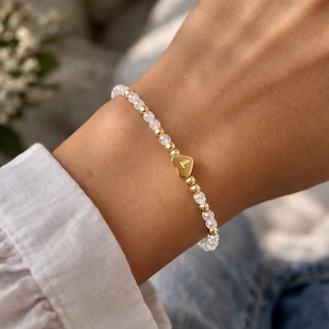May include: Gold beaded bracelet with clear crystal beads and a gold heart charm with the letter "T". The bracelet is on a wrist, and the background is blurred.