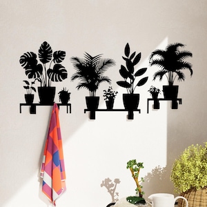 May include: Black metal wall-mounted shelf with five hooks, featuring a silhouette design of potted plants. A colorful towel hangs from one of the hooks.