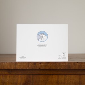 May include: A white greeting card with a blue and white mountain logo that says "Lakeland Explorer". The card also has text that reads "Ullswater and Hallin Fell" and "&copy; Christian Shaw 2020". The card is on a wooden surface.