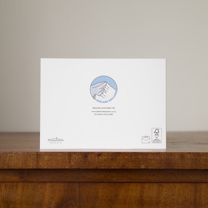 May include: A white greeting card with a blue and white mountain logo that says "Lakeland Explorer". The card also has text that reads "Ullswater and Hallin Fell" and "&copy; Christian Shaw 2020". The card is on a wooden surface.