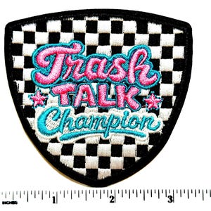 May include: Embroidered patch with a black border and a checkered background. The patch features the words "Trash Talk Champion" in pink and teal lettering, with star accents. The patch is shield-shaped.