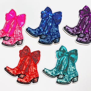 May include: Five colorful patches of cowboy boots with large sequined bows. The boots are in shades of pink, blue, purple, red, and teal. Each boot has a floral pattern and a small heel. The bows are a contrasting color to the boots.