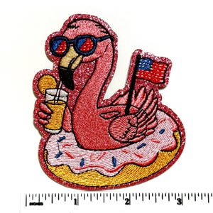 May include: Embroidered patch featuring a pink flamingo wearing sunglasses and floating on a donut-shaped float. The flamingo holds a drink and a small American flag. The patch has glitter accents.