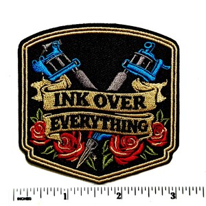 May include: A shield-shaped embroidered patch featuring two crossed blue tattoo machines above a gold banner with the black text INK OVER EVERYTHING and red roses with green leaves at the bottom, positioned above a ruler showing 4 inches in width.