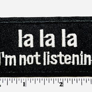 May include: Black rectangular patch with white embroidered text that reads "la la la I'm not listening." The patch is approximately 3 inches wide and has a black border.