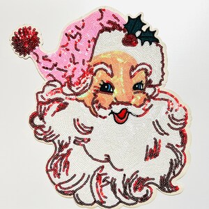 May include: A sequined Santa Claus face with a pink hat, white beard, and rosy cheeks. The Santa Claus face is decorated with red sequins and has blue eyes and a red mouth. A sprig of holly is on the hat.