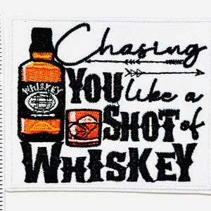 May include: Embroidered patch with the text "Chasing You like a Shot of Whiskey." The design includes a whiskey bottle and a glass of whiskey, all in shades of orange, black, and white. The patch is rectangular and has a white background.