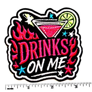 May include: An embroidered patch featuring a martini glass with a lime slice and pink liquid, surrounded by pink flames and blue stars. The text reads DRINKS ON ME in white and pink lettering. A ruler shows the patch is approximately 3.5 inches wide.