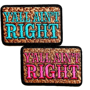 May include: Two rectangular patches with the text "Y'ALL AIN'T RIGHT" embroidered in turquoise and pink thread. The patches have a brown, textured background and a black border. The patches are likely for clothing or accessories.