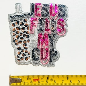 May include: A sequined patch with the phrase "JESUS FILLS MY CUP" in pink, black, and silver. The patch includes a leopard print cup design. A yellow tape measure is placed below the patch.