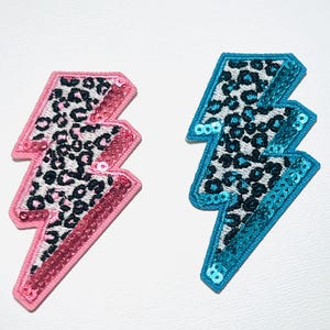 May include: Two embroidered lightning bolt patches. One is pink with a leopard print design and pink sequins. The other is blue with a leopard print design and blue sequins. Both are on a white background.