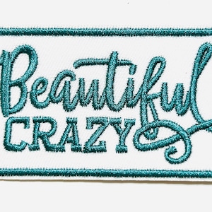 May include: A rectangular white patch with teal embroidered text that reads "Beautiful Crazy." The patch has a teal border and is approximately 2 inches in height. The text is in a cursive font.