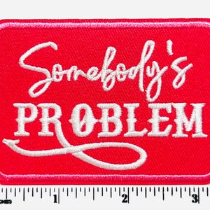 May include: A rectangular red patch with white embroidered text that reads "Somebody's PROBLEM". The patch has a white border and a ruler at the bottom, measuring in inches. The patch is approximately 3 inches wide.