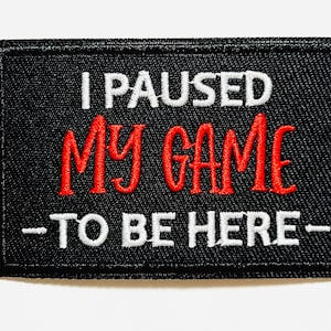 May include: Black rectangular patch with white and red embroidered text. The text reads "I PAUSED MY GAME TO BE HERE". The patch is approximately 2 inches wide and 1 inch tall.