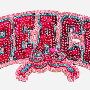 May include: A pink sequined patch with the word "BEACH" in red letters outlined in teal. A teal and pink bow is at the bottom. The patch is approximately 2 inches wide.