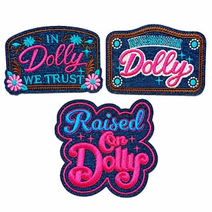 May include: Three embroidered patches on a dark blue denim-like background. One patch reads IN DOLLY WE TRUST in pink and blue. Another reads RAISED ON DOLLY in pink and blue. The third patch reads Raised On Dolly in pink and blue script.
