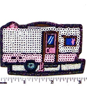 May include: A camper trailer patch featuring a dark purple glitter background, white and light pink sequins, a brown door with a blue window, and a blue wheel. A ruler placed below the patch shows it measures approximately 3.5 inches in width.