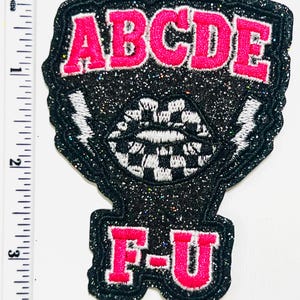 May include: Embroidered patch with the letters ABCDE in pink, outlined in black with a glittery texture. Below, the letters F-U are also in pink. The patch features a checkered mouth design and lightning bolts.