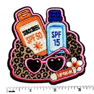 May include: An embroidered patch featuring an orange sunscreen bottle labeled SPF 50, a blue sunscreen bottle labeled SPF 15, heart-shaped sunglasses, and a lip balm tube with a flower, all on a leopard print background, next to a 3-inch ruler.