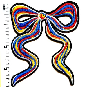 May include: Embroidered bow patch with a black border. The bow features blue, red, yellow, and white stripes. The patch measures approximately 3 inches in height.