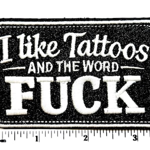 May include: A rectangular black embroidered patch with white stitched borders and white text that reads I like Tattoos AND THE WORD FUCK. A white ruler is positioned along the bottom edge, showing the patch is approximately 3.5 inches wide.