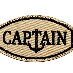 May include: Tan embroidered patch with the word "CAPTAIN" in black, featuring an anchor. The patch is approximately 2 inches long and 1 inch tall, with a black border and decorative stitching.