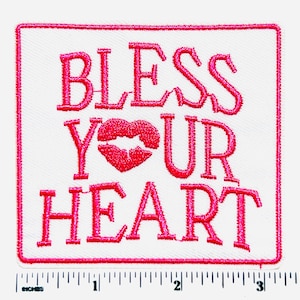 May include: Square white patch with a pink border and the embroidered phrase "BLESS YOUR HEART" in pink. The word "YOUR" features a red lip design in place of the letter "O".