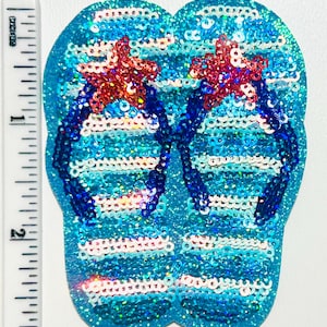 May include: A sequined flip-flop design in shades of blue, white, and red. The flip-flops have a turquoise base with white horizontal stripes. The straps are dark blue, and the design includes two red starfish. The design is approximately 3 inches in length.