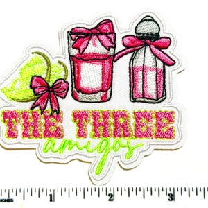 May include: Embroidered patch featuring a cocktail glass, a bottle, and a lime with a pink bow. The text "THE THREE amigos" is in pink and green. The patch is on a white background.