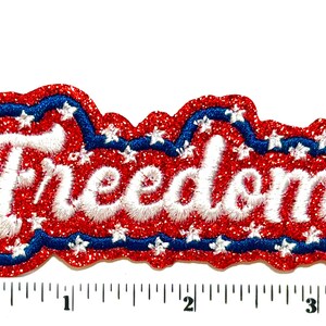 May include: A red glittery embroidered patch with the word Freedom in white script lettering. The patch features a blue border and small white stars, measuring approximately 4 inches in length as shown by the visible ruler at the bottom.