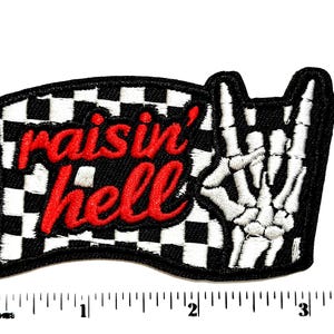 May include: An embroidered patch featuring a black and white checkered flag background with the red text "raisin' hell" and a white skeleton hand making a rock and roll gesture. A ruler shows the patch is approximately 3.5 inches wide.