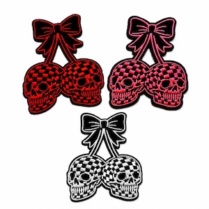 May include: Three embroidered patches featuring a pair of skulls connected by a bow. Two patches have red and black checkered skulls with black bows, while one patch has white and black checkered skulls with a black bow, all with black borders.