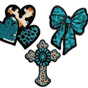 May include: A collection of embroidered patches featuring a turquoise bow, a cross, and three hearts. The hearts have a turquoise, leopard print, and cowhide pattern. The bow and cross have turquoise and leopard print patterns.