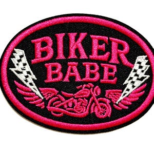May include: Oval-shaped patch with the words "BIKER BABE" in bright pink, outlined in black and a pink border. The design includes a motorcycle and lightning bolts with wings. The patch measures approximately 2 inches in height.