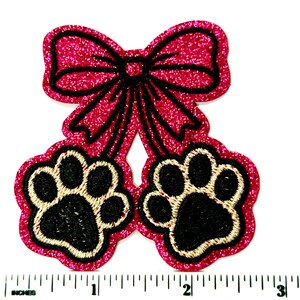 May include: A pink glittery patch featuring a bow and two paw prints. The bow and paw print outlines are black, with the paw pads in black and gold. The patch is approximately 3 inches wide.